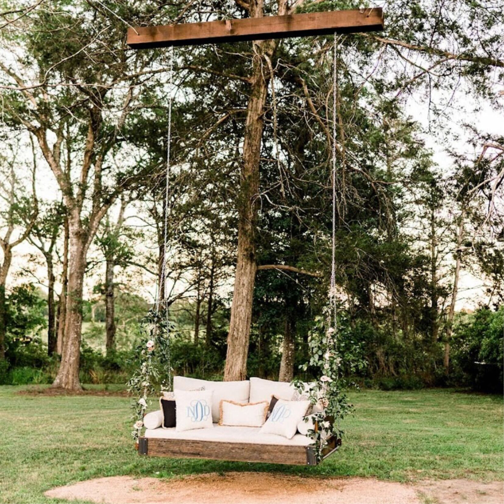 A rustic wooden swing suspended by ropes, with a cushioned seat and pillows, set in a grassy outdoor area surrounded by trees.