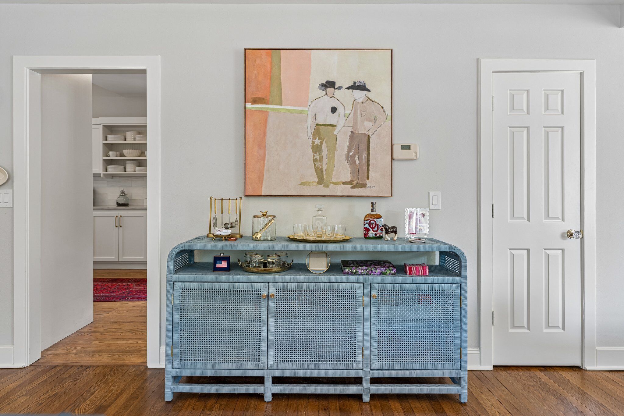 A room with a blue wicker console table, a painting above it, and a white door to the right.