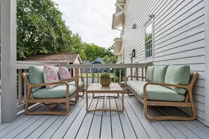 Una terraza de madera con dos sillones de mimbre y una mesa, una pequeña planta en maceta, y una casa al fondo.