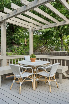 Un patio techado al aire libre con una mesa redonda y sillas de mimbre, rodeado por una terraza de madera.