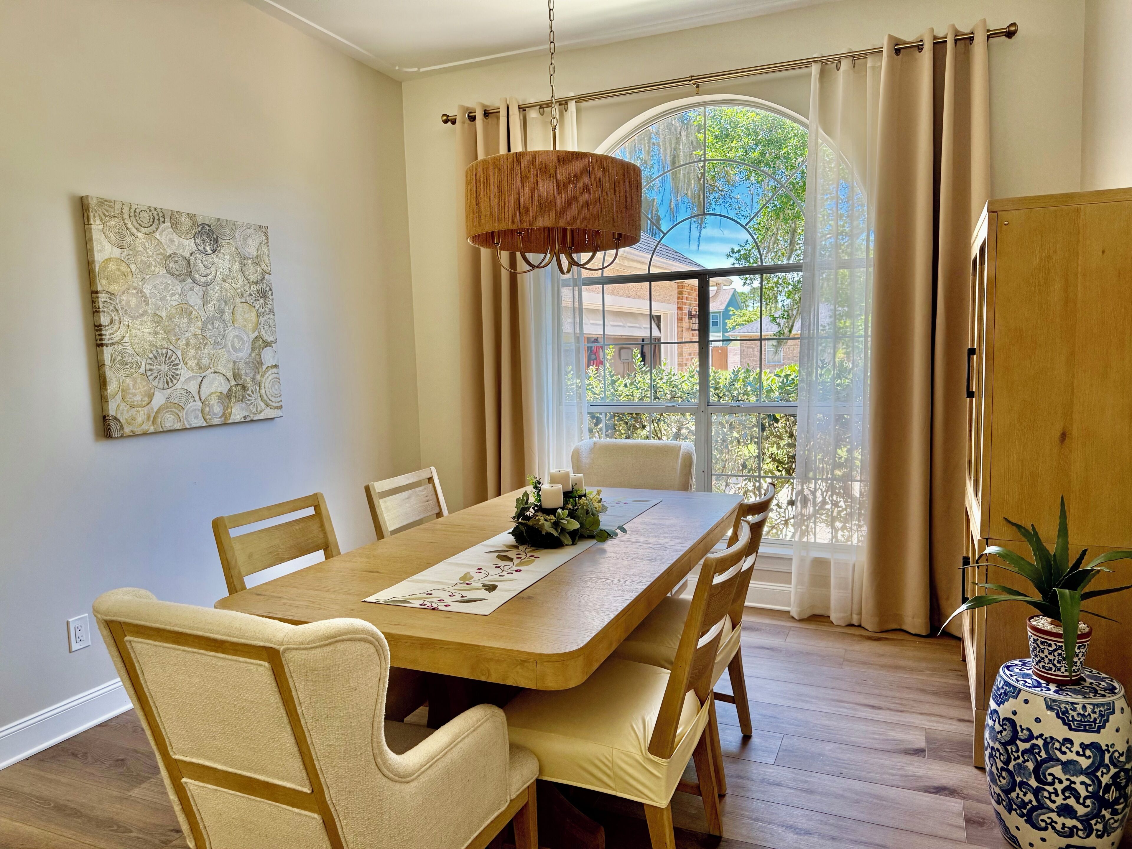 A dining area with a wooden table, cushioned chairs, a hanging light fixture, a window with curtains, and a vase with a plant.