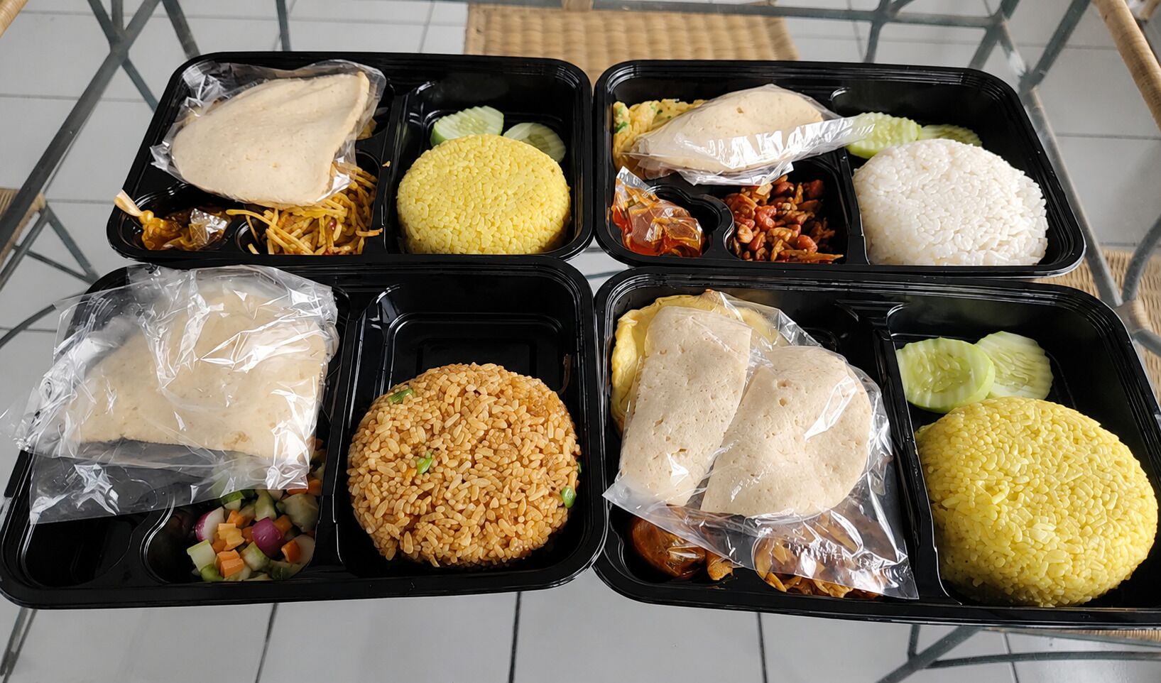 Cuatro recipientes negros con comida, incluyendo arroz, verduras y carne, sobre una mesa.