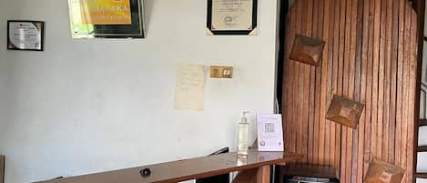 Una recepción de hotel con un mostrador de madera, un cubo de basura negro con etiqueta de almacenamiento de agua, y varios certificados y reconocimientos en la pared.