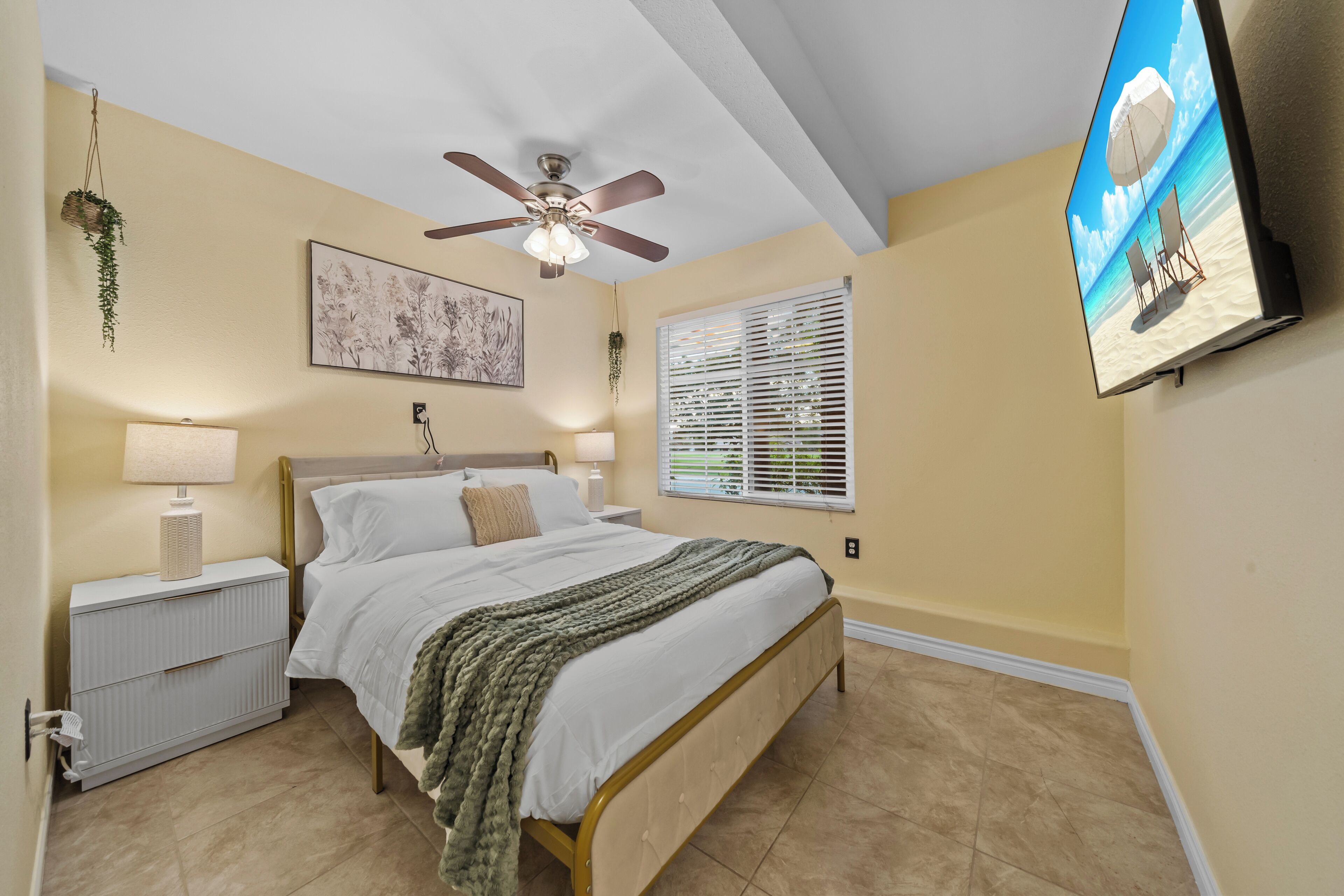 A bedroom with a bed, bedside tables, a ceiling fan, a wall-mounted TV, and a window with blinds.