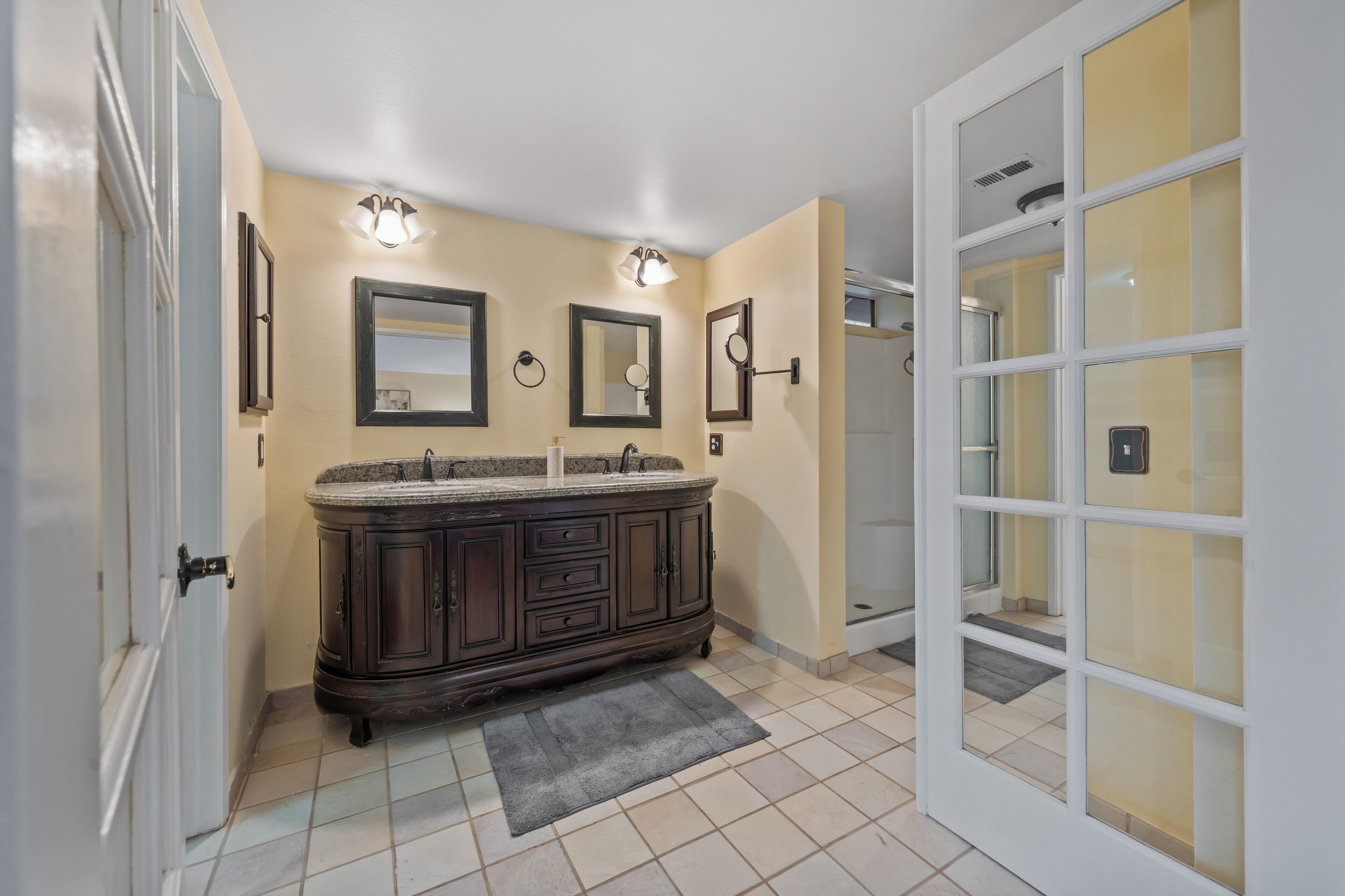 Une salle de bain avec un meuble-lavabo en bois foncé, deux miroirs fixés au mur et une cabine de douche en verre.