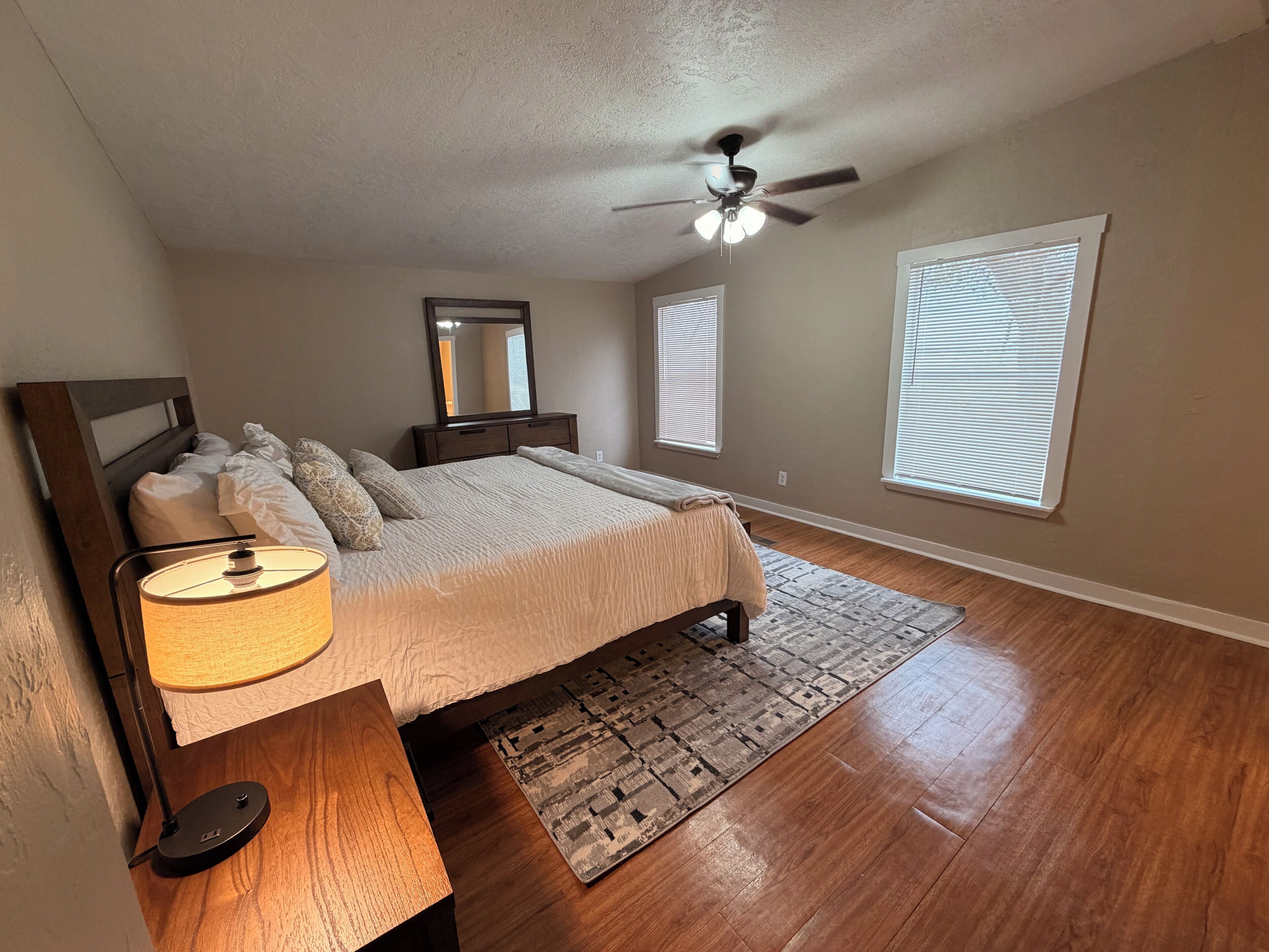 A bedroom with a bed, bedside table, lamp, ceiling fan, windows, and a rug.