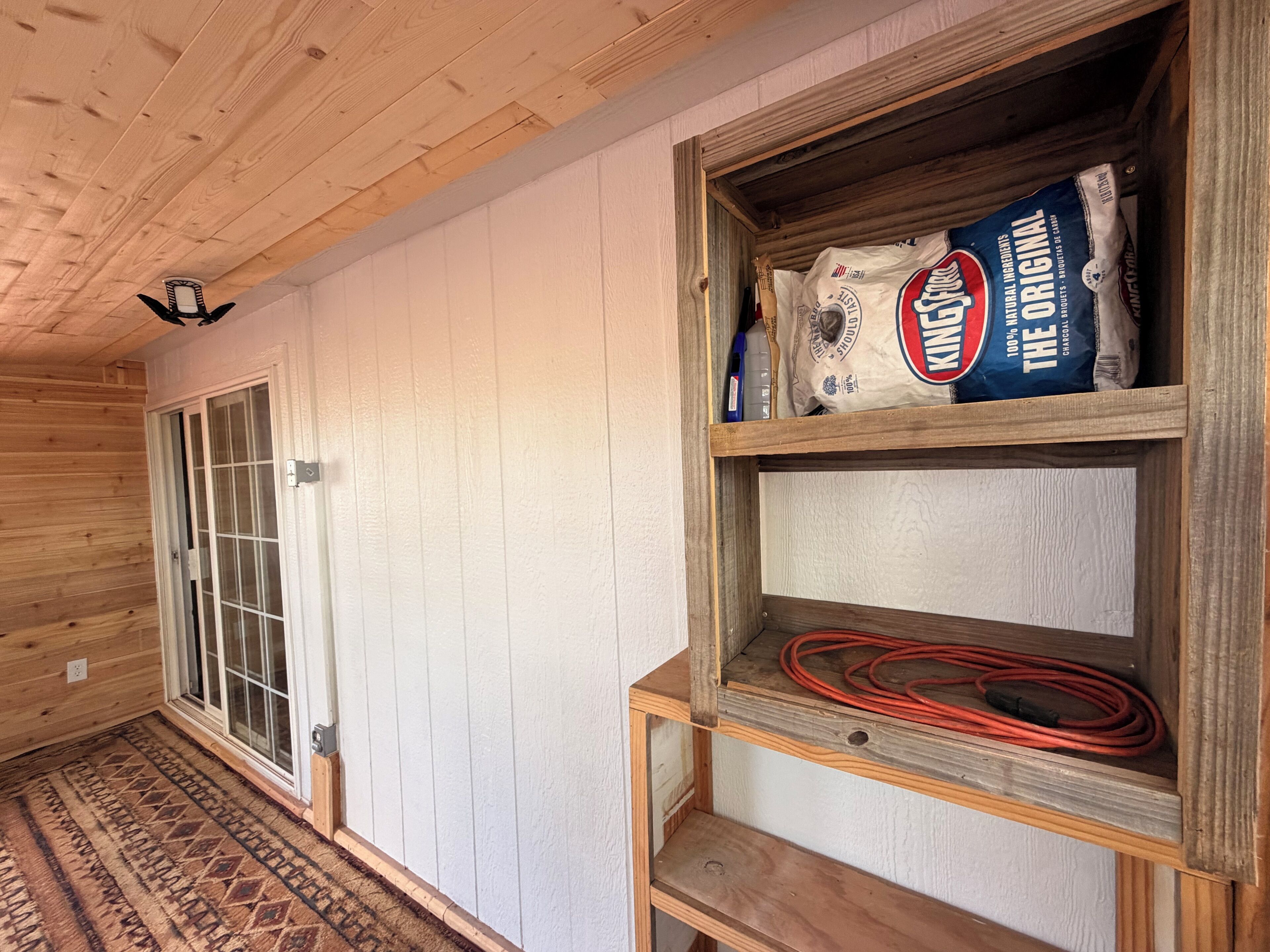 Una stanza con soffitto in legno, una porta con pannelli di vetro, una mensola con un sacco di carbone e un cavo di prolunga rosso.