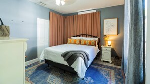 A bedroom with a bed, a nightstand with a lamp, a window with curtains, and a wall-mounted artwork. - El Apartemento | Cozy 1BR Cottage with Private Deck Near Ybor City, Armature Works & Downtown Tampa (Tampa)