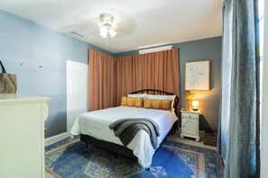 Un dormitorio con cama, mesita de noche con lámpara, ventana con cortinas y una obra de arte colgada en la pared.
