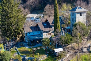 Uma casa grande, de vários andares, com uma torre de pedra, rodeada por árvores e um jardim bem cuidado.