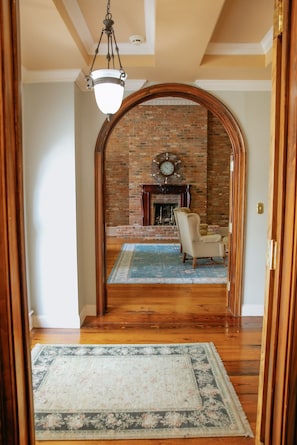 A hallway leading to a living room with a brick fireplace, a clock, and a chair. - French Quarter-Style Retreat, Private Pool, Theater, Wine Cellar & Game Room (Lafayette)