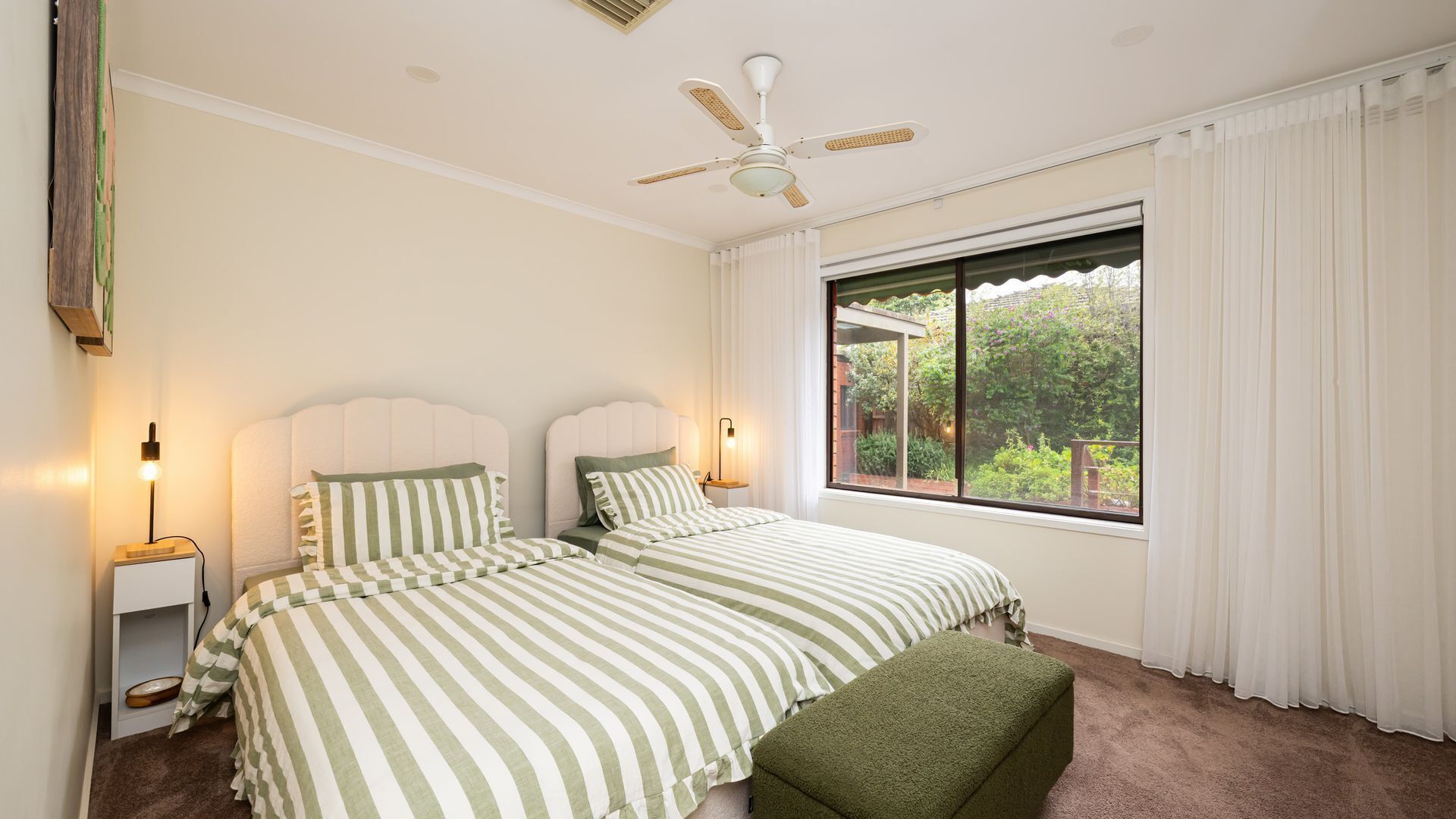 A bedroom with two beds, a ceiling fan, bedside lamps, a window with curtains, and a view of greenery outside.
