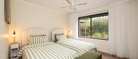 A bedroom with two beds, a ceiling fan, bedside lamps, a window with curtains, and a view of greenery outside.