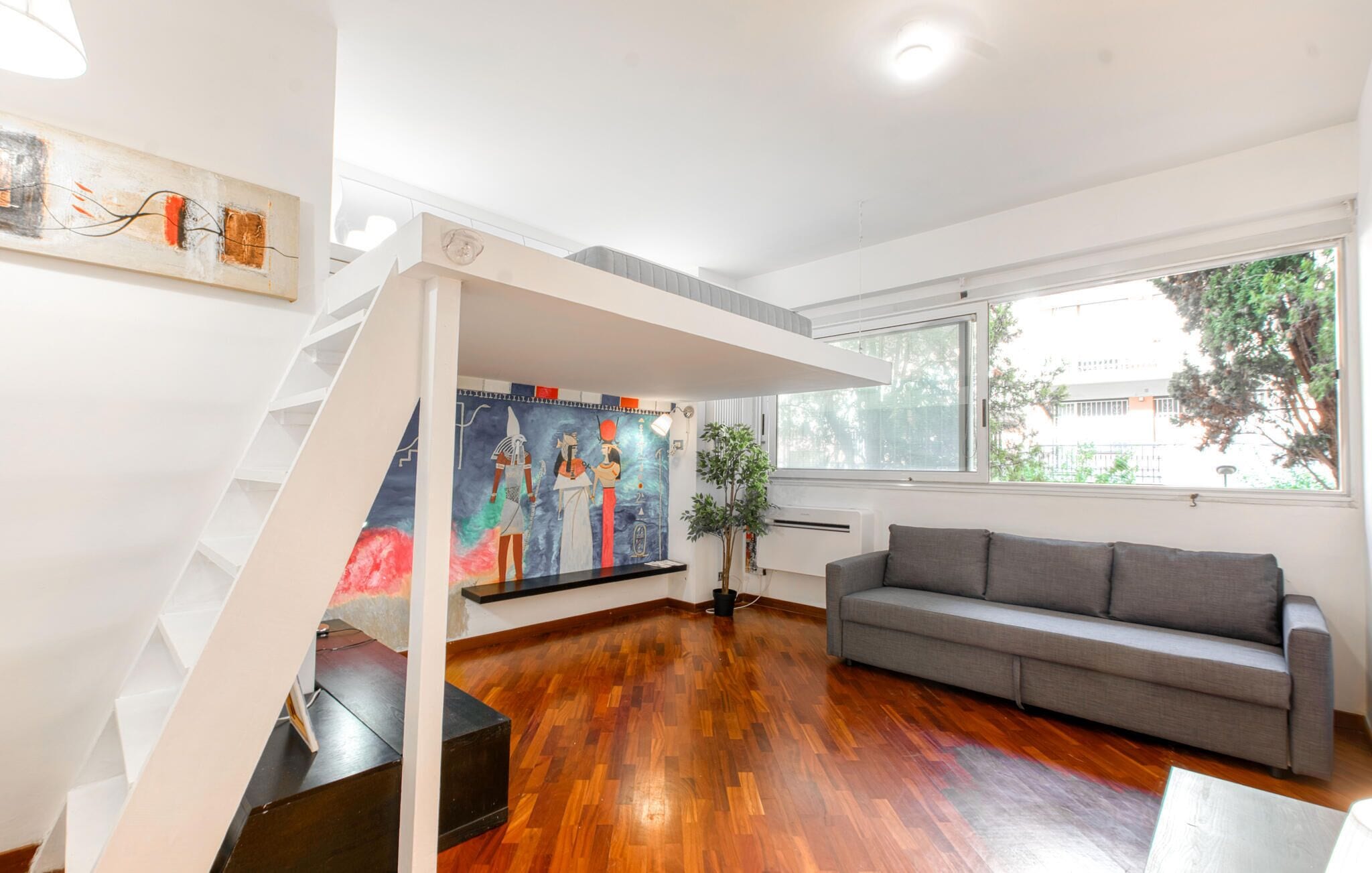 Un salon moderne avec un plancher en bois, un canapé gris, un escalier doté d’une rampe blanche et une grande fenêtre donnant sur la forêt.
