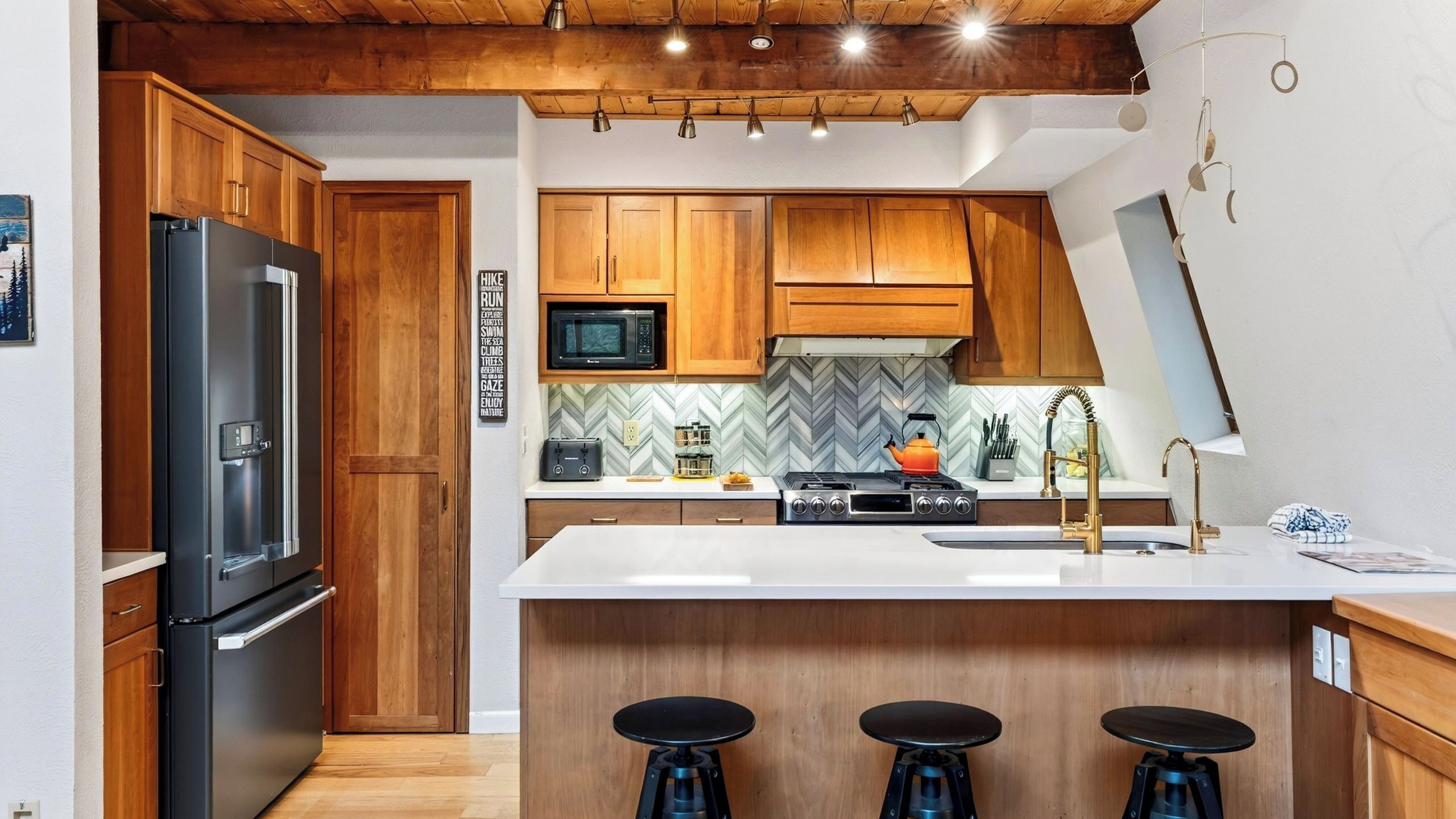 Una cocina moderna con armarios de madera, un refrigerador de acero inoxidable, un microondas, una estufa a gas y un lavamanos con grifo dorado. Hay tres taburetes en la isla.
