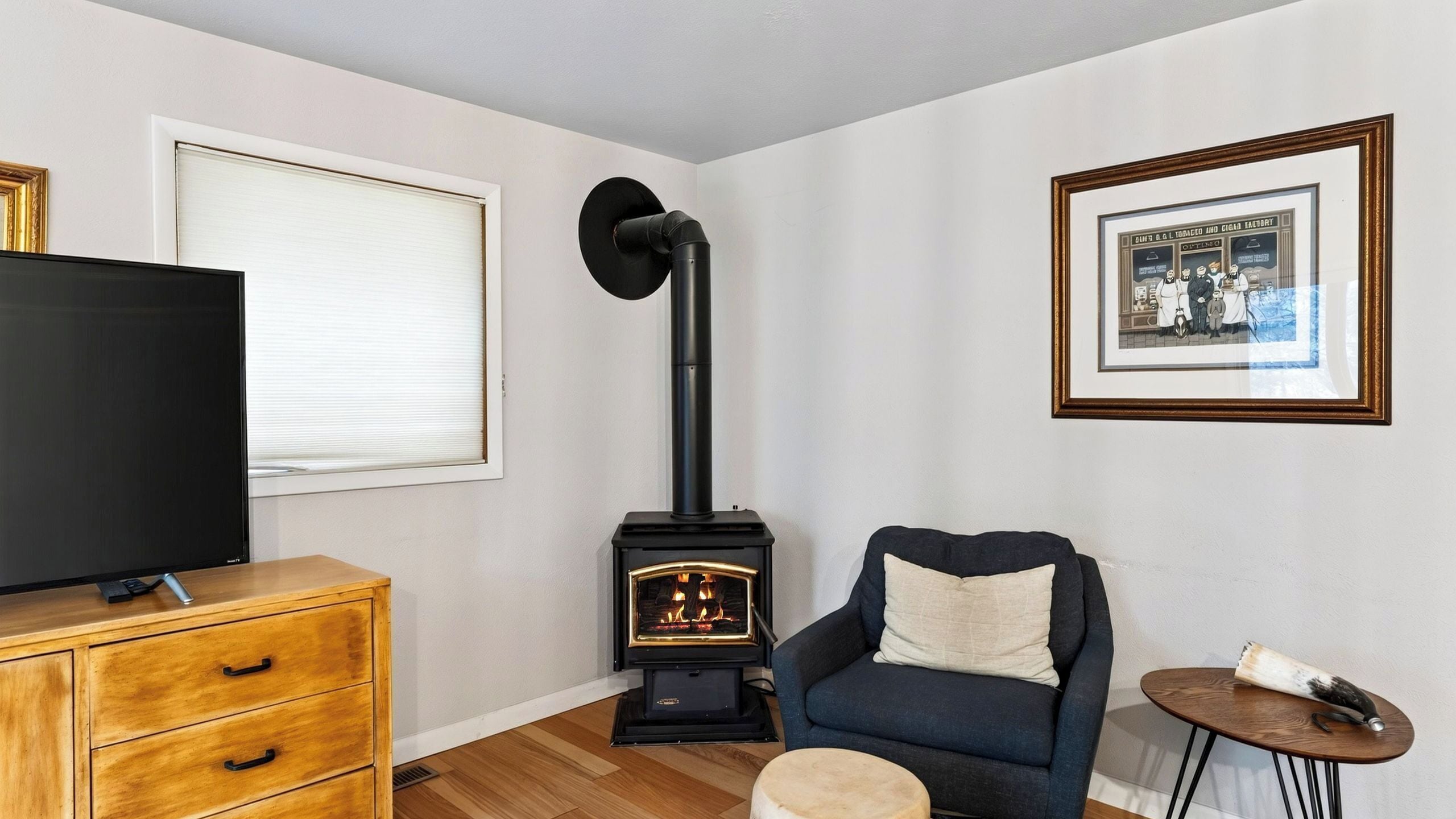 A living room with a television, a wooden cabinet, a blue armchair, a small round table, and a black wood-burning stove.