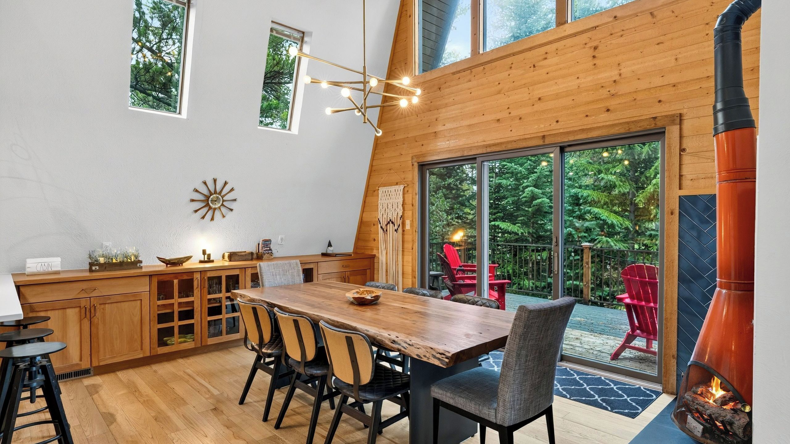 Interior de una cabaña con una mesa de comedor de madera, sillas, una cocina y un gran ventanal corredero que da a una terraza exterior con sillas rojas y un estufa de leña.