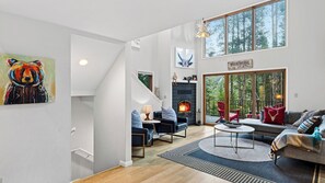 A modern living room with a fireplace, seating area, and a large window overlooking a forest. - Pitch & Pine by Avantstay Hot Tub + Ping Pong + Arcade + Loft Lounge Ski, Hike + Main Street (Breckenridge)