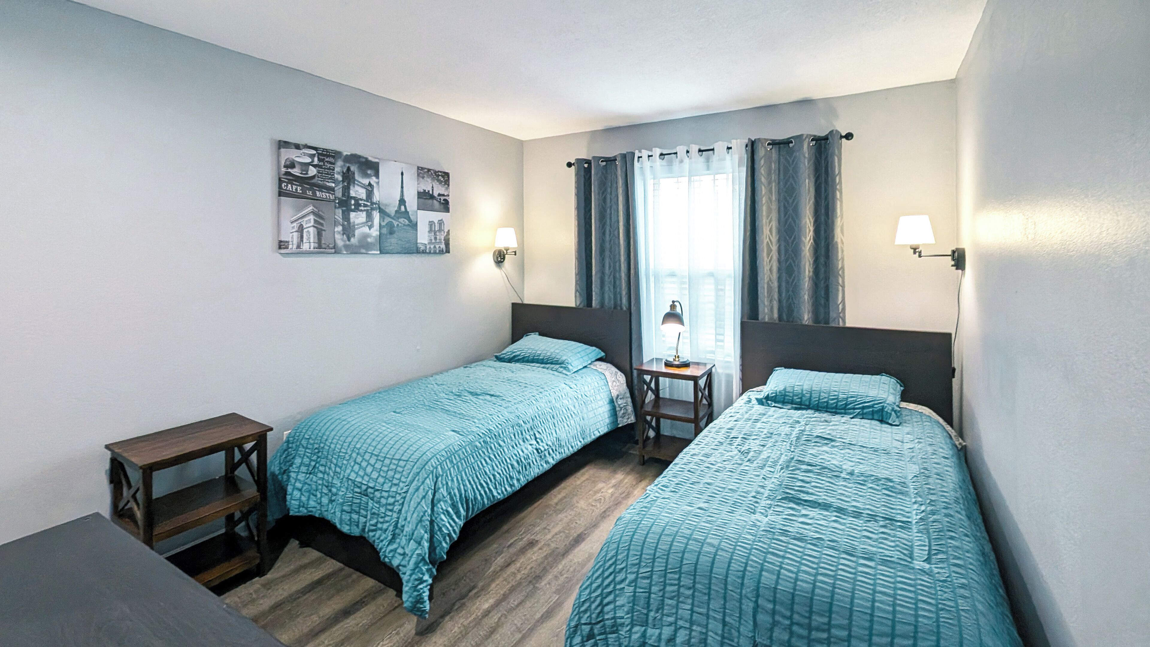 Quarto de hotel com duas camas de solteiro, uma mesa pequena, uma cadeira, mesa de cabeceira e luminária na parede.