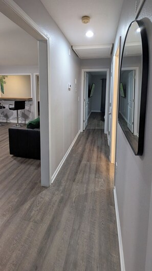 A hallway with a wooden floor, a mirror on the wall, and a door leading to another room.