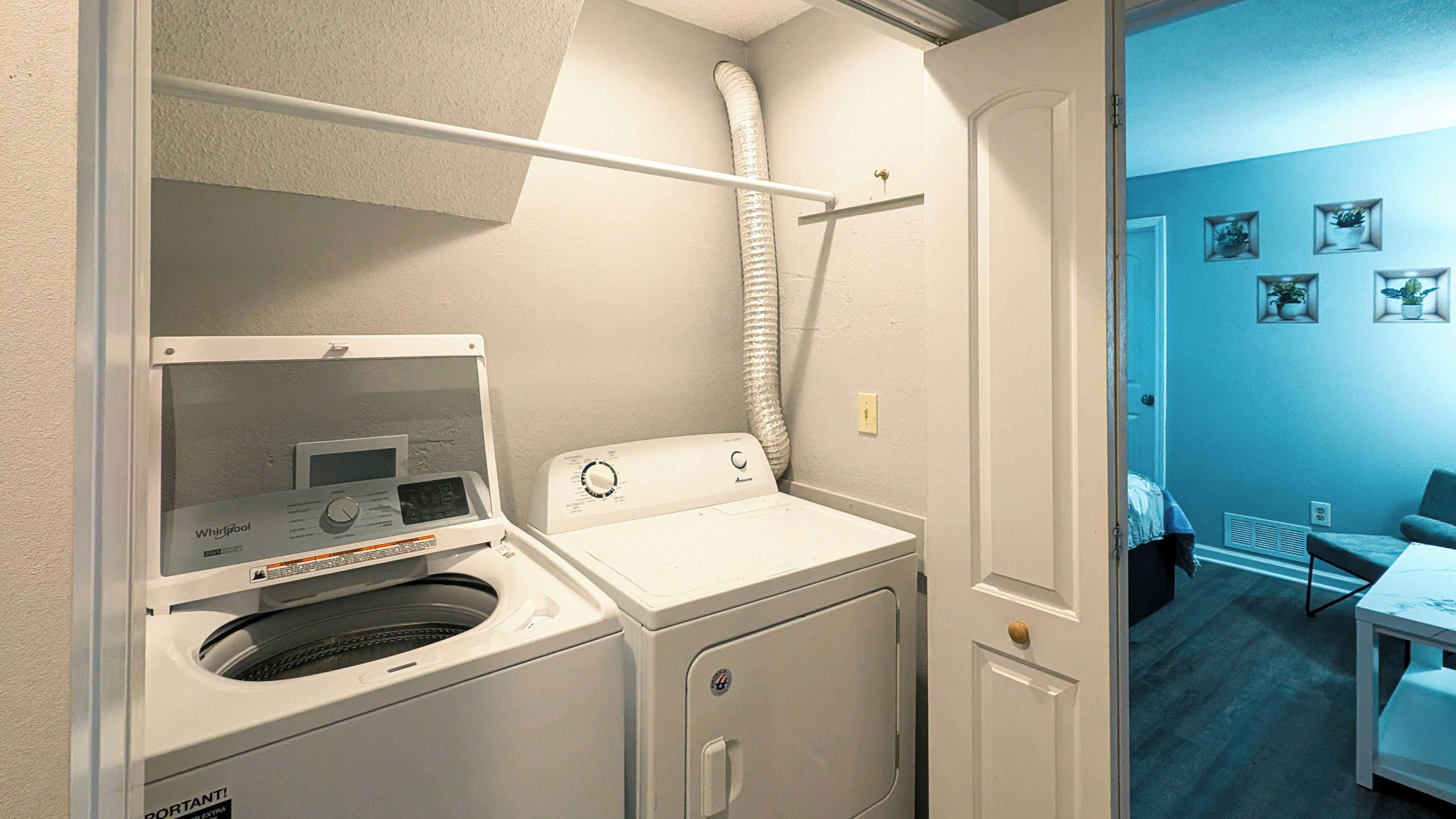 A laundry room with a front-loading washer and dryer, a vent hose, and a door leading to a room with a blue wall and a table.