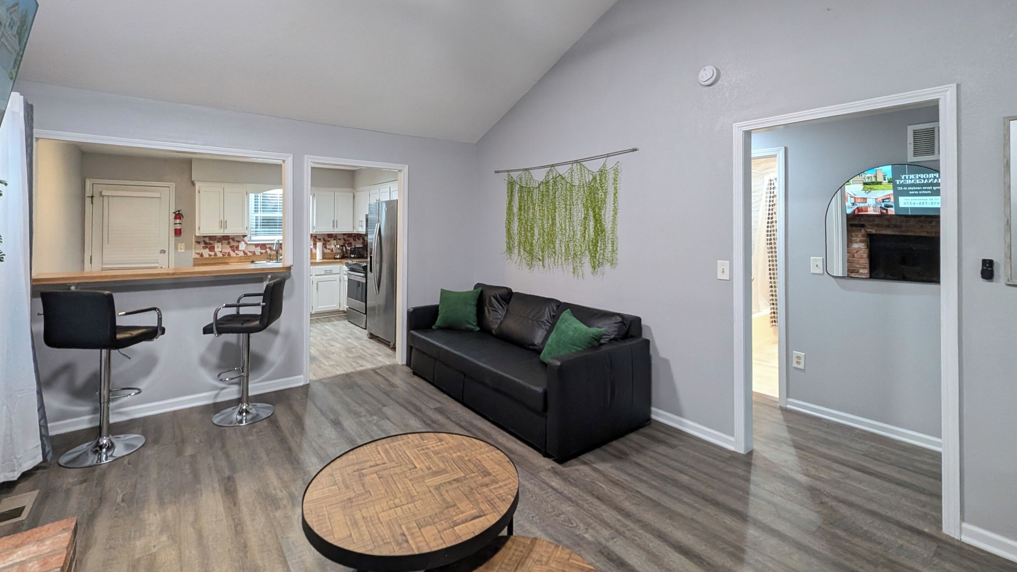 Un salon moderne avec un canapé noir, deux coussins verts, une table basse ronde en bois et une cuisine visible à travers une porte ouverte.