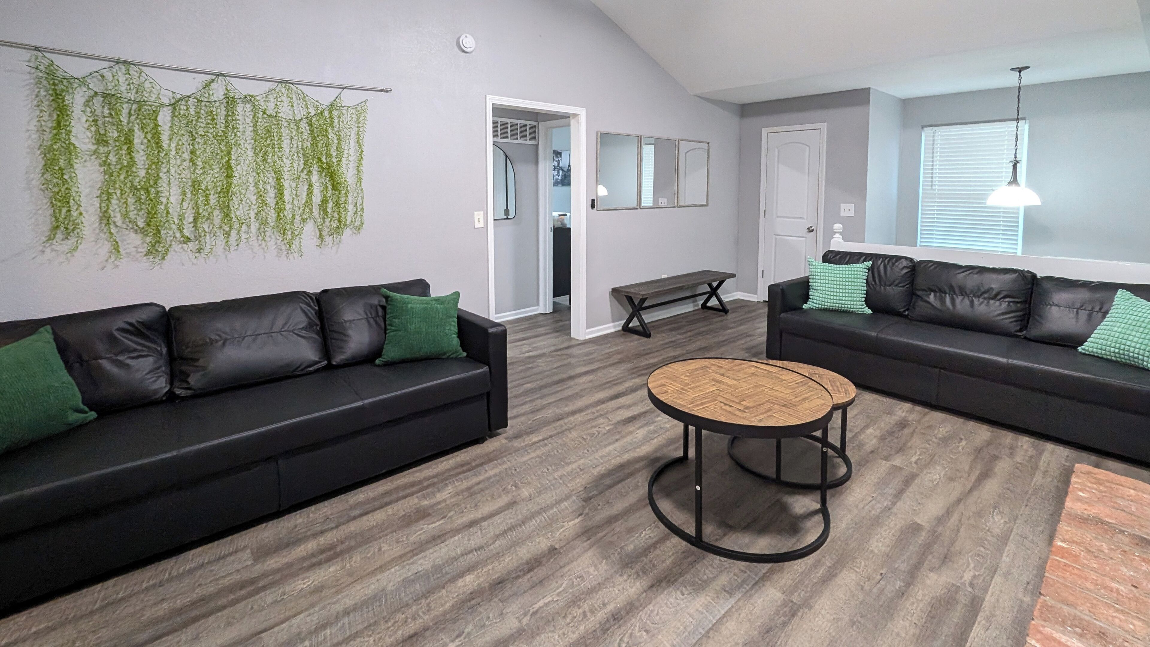 A modern living room with two black leather sofas, a round wooden coffee table, and a hanging green plant.
