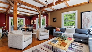 A spacious living room with high ceilings, wooden beams, a dining area with chairs and a table, and large windows with a view of greenery. - Bow & Beam by Avantstay Timber-frame Retreat + Chef's Kitchen + Game Room 3.5 Acres + Trails (Sheffield)