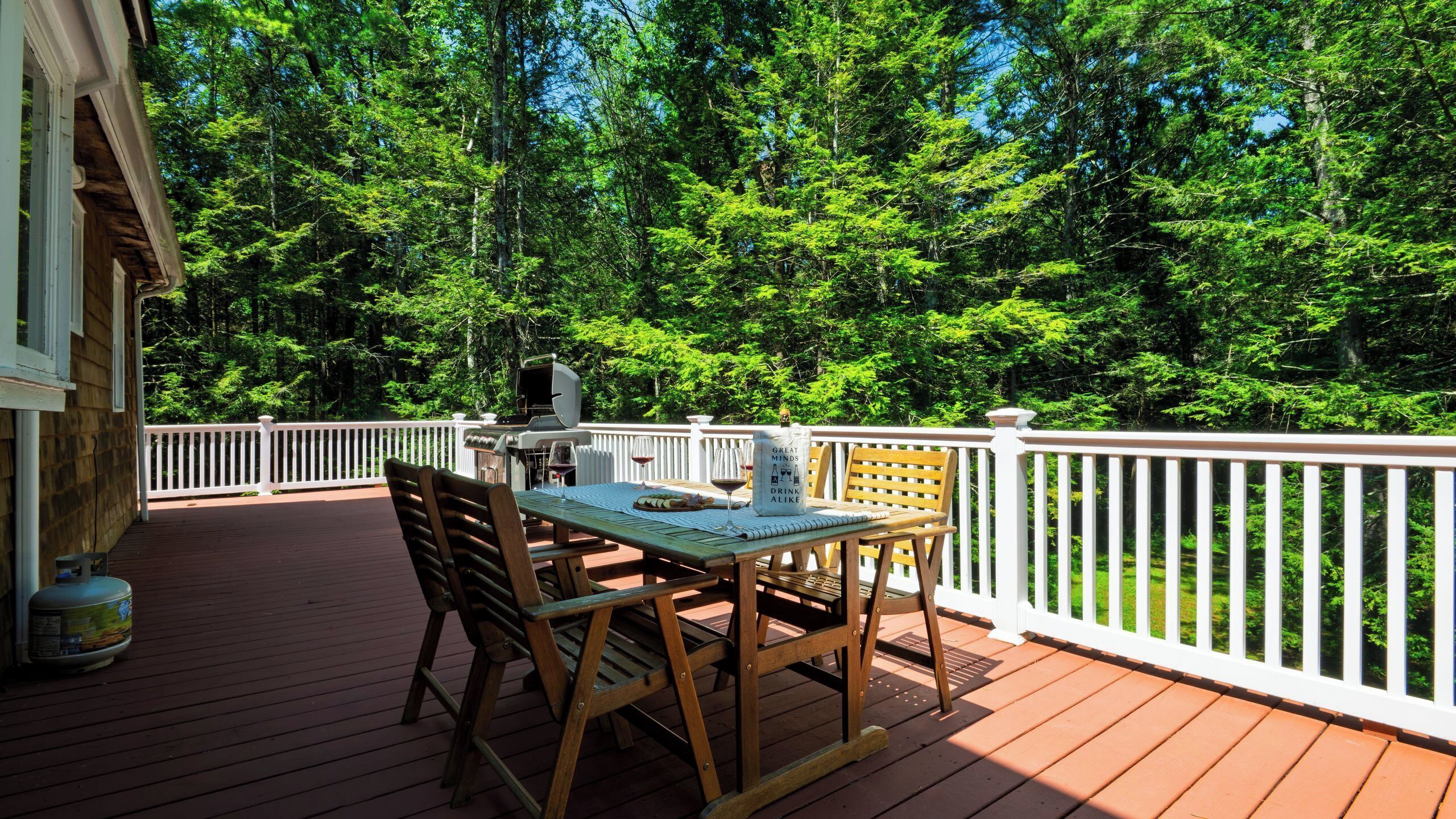 A wooden deck with a table and chairs, surrounded by a white railing, overlooking a forested area.