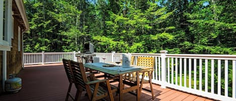 A wooden deck with a table and chairs, surrounded by a white railing, overlooking a forested area.