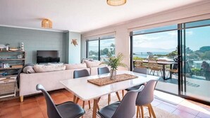 A modern living room with a sofa, dining table, chairs, and a TV. There is a balcony with a view of the landscape. - The Rooftop - Bicheno Accommodation (Bicheno)