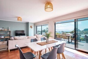 Ein modernes Wohnzimmer mit einer Couch, einem Esstisch, Stühlen und einem Fernseher. Ein Balkon bietet Blick auf die Landschaft.