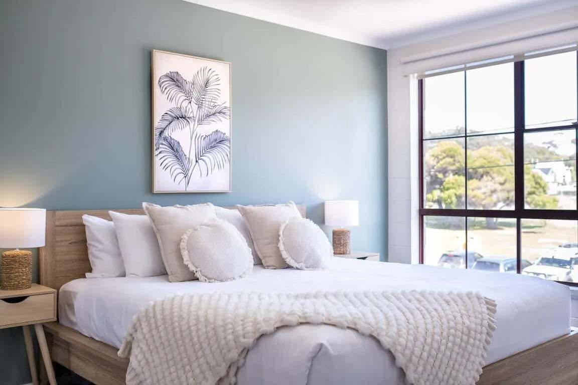 Quarto com cama, travesseiros, abajur, uma obra de arte na parede e janela com vista.