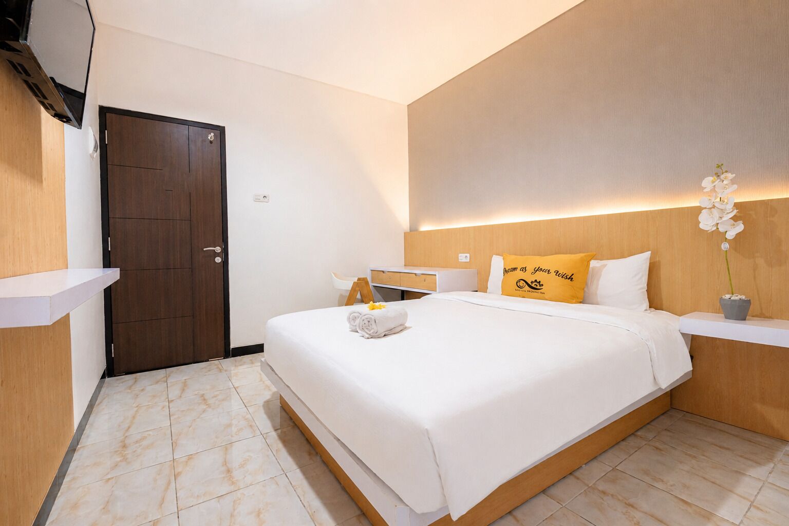 A hotel room with a bed, a bedside table, a nightstand, a door, and a small potted plant.