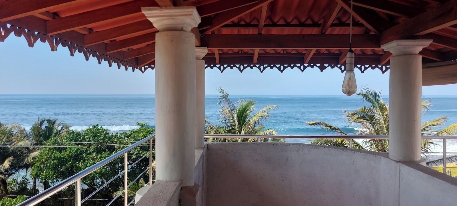 A covered balcony with a view of the ocean, palm trees, and a clear sky.