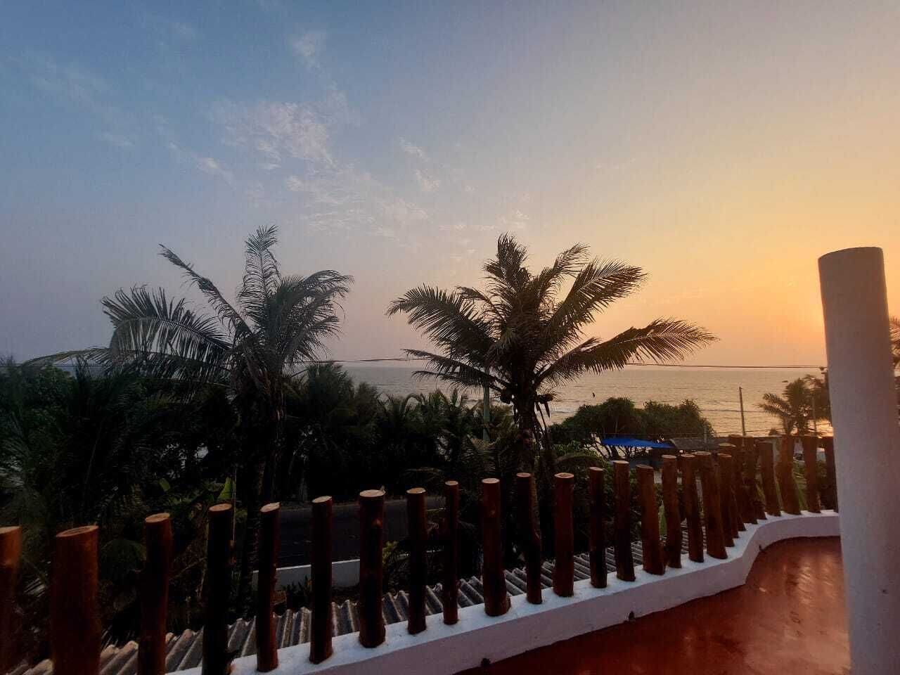 Un balcon avec vue sur la mer au coucher du soleil, des palmiers et une balustrade blanche.