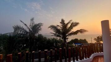 Un balcon avec vue sur la mer au coucher du soleil, des palmiers et une balustrade blanche.