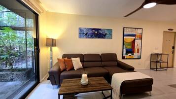 A modern living room with a sofa, a coffee table, and a lamp. There are abstract paintings on the wall and a view of greenery outside.
