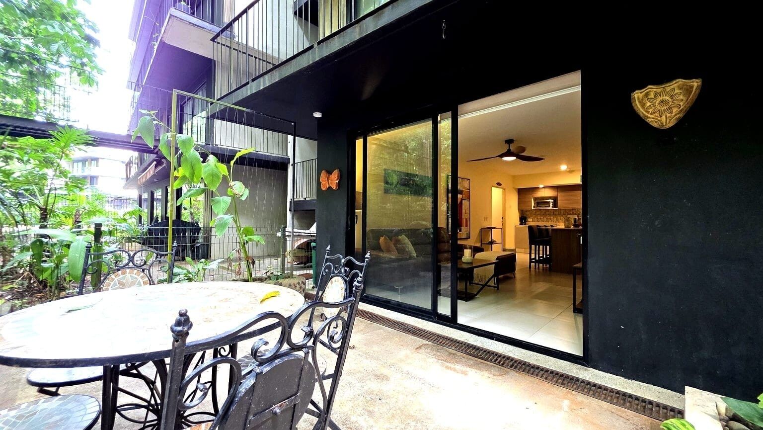 A patio with a round table and chairs, a sliding glass door leading to an interior space with a ceiling fan, and a decorative wall plaque.