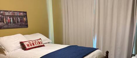 A bedroom with a bed, a ceiling fan, a wall with a fishing lure poster, and a window with curtains.
