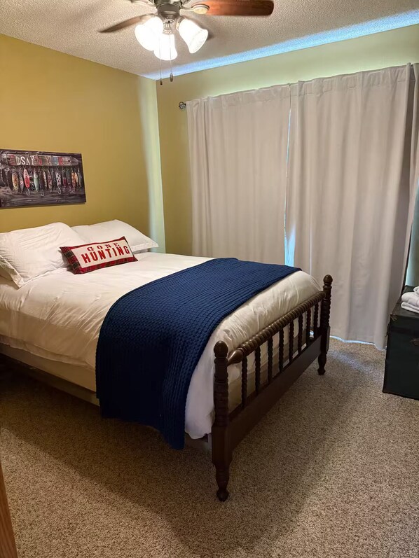 A bedroom with a bed, a ceiling fan, a wall with a fishing lure poster, and a window with curtains.