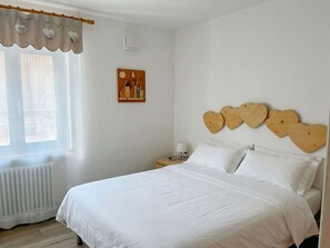 Ein Schlafzimmer mit einem Bett, einem Nachttisch, einer Lampe, einem Heizkörper, einem Fenster mit Vorhängen und einem Wandbild.