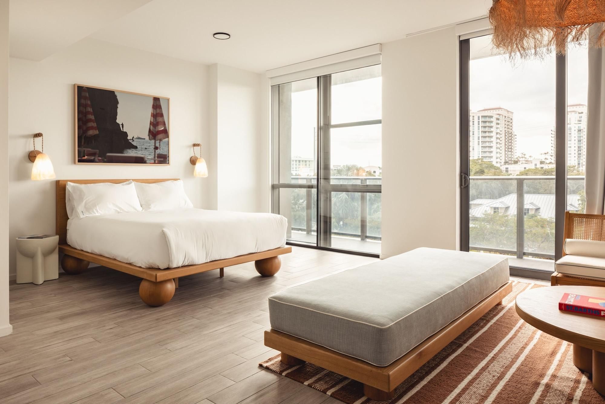 Habitación de hotel moderna con una cama grande, un sofá más pequeño, una mesa de comedor y vistas a la ciudad a través de amplios ventanales.