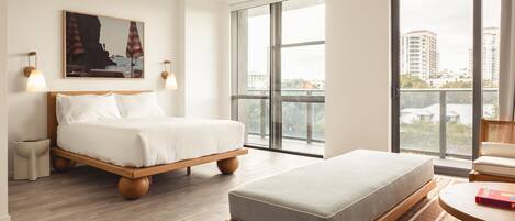 Habitación de hotel moderna con una cama grande, un sofá más pequeño, una mesa de comedor y vistas a la ciudad a través de amplios ventanales.