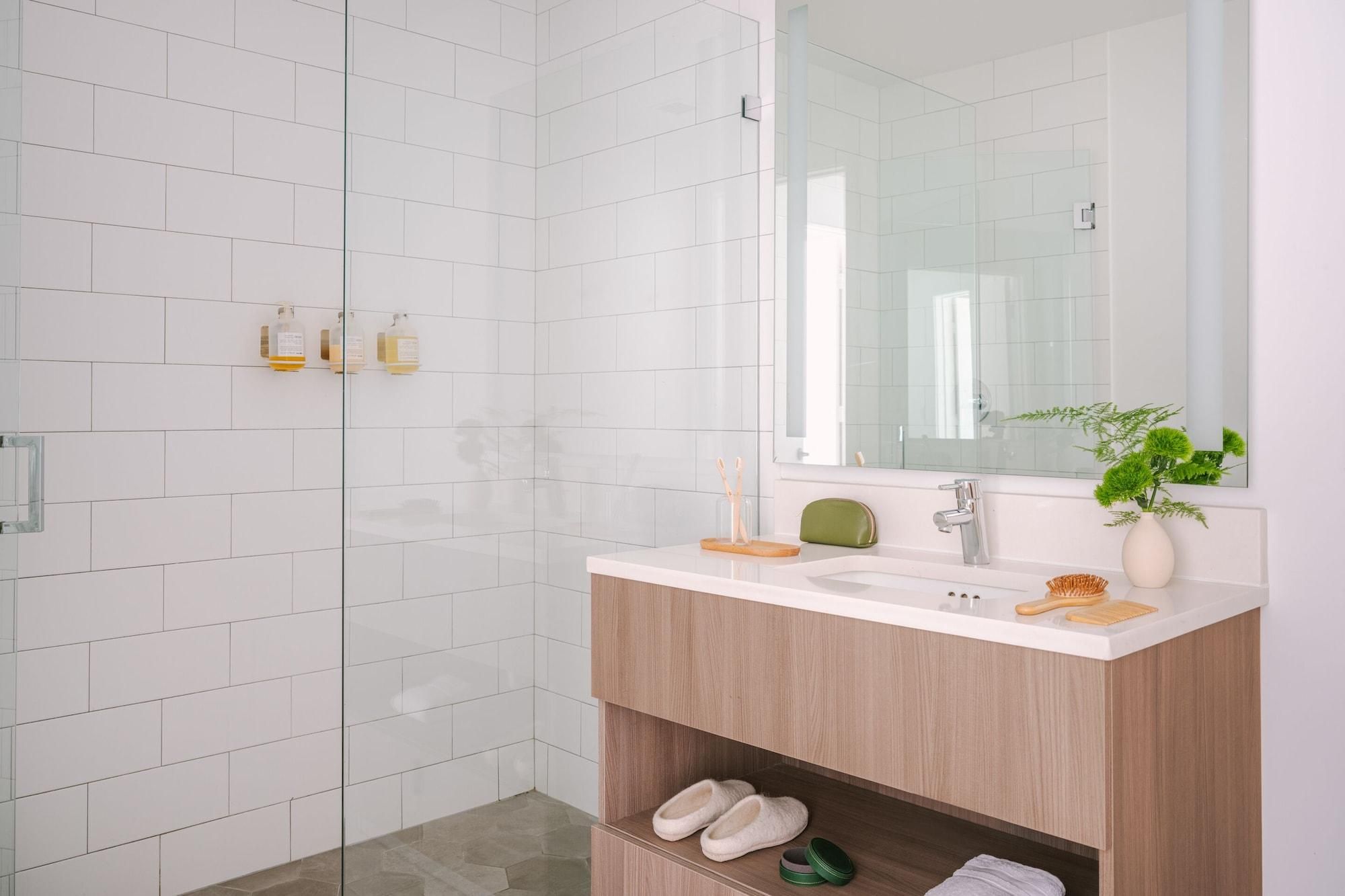 A modern bathroom with white tiles, a glass shower enclosure, a wooden vanity, and a mirror.