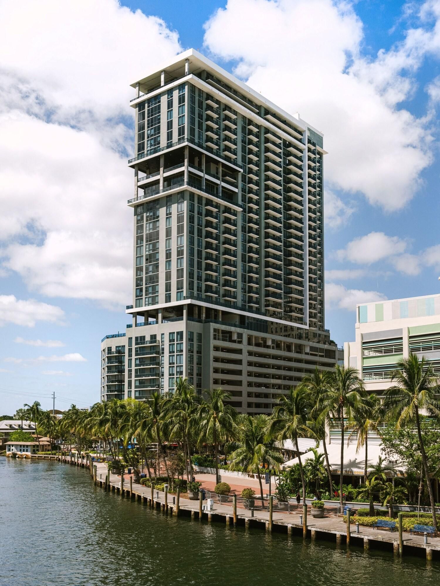 A modern high-rise hotel with multiple balconies, a waterfront view, and a palm-lined promenade.