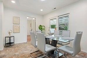 A modern dining area with a glass table and metal chairs, a small side table with a sculpture, and a large window with a view of greenery. - Casa La Vuelta - 4 King Suites Near Miramar Beach (Montecito)