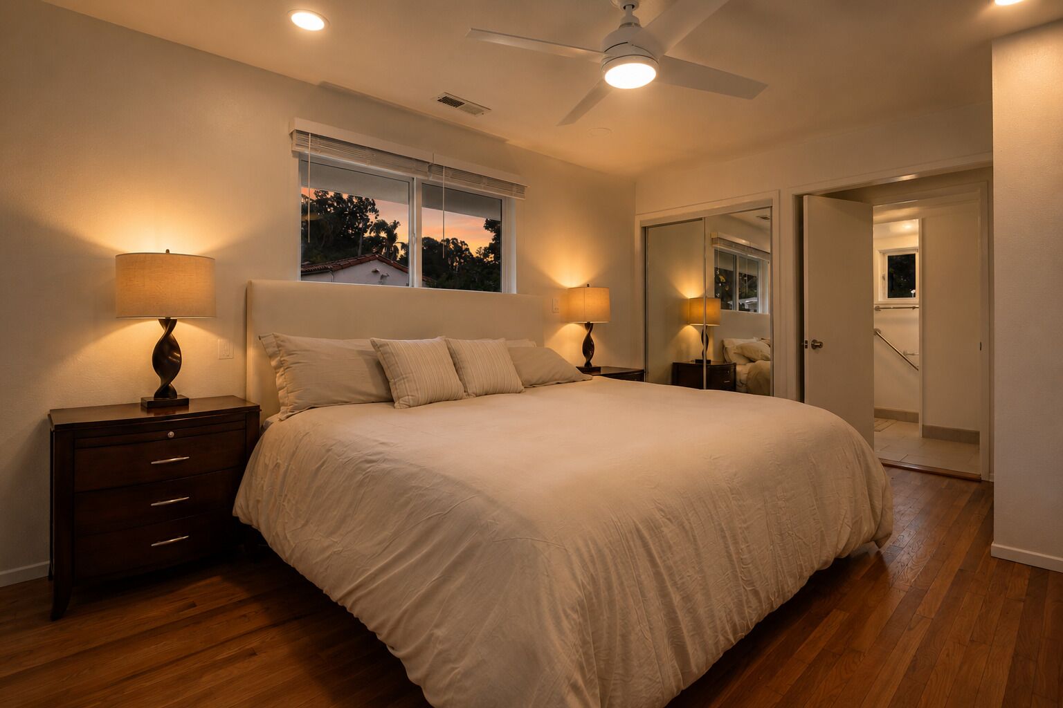 A bedroom with a large bed, two bedside tables with lamps, a ceiling fan, and a window with blinds.