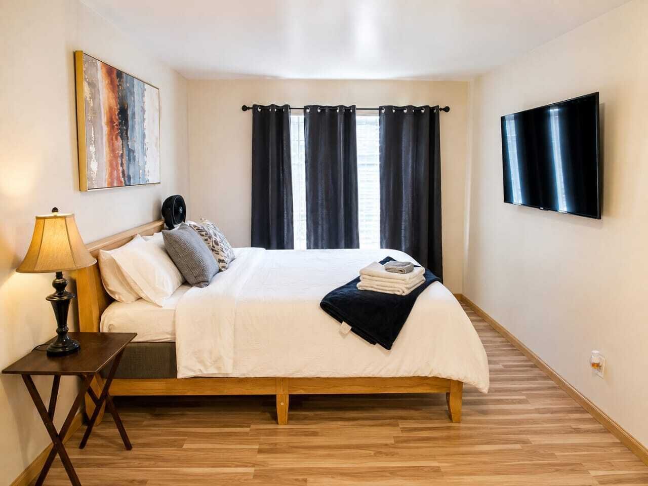 Un dormitorio con cama, mesita de noche con lámpara, televisor montado en la pared y ventana con cortinas.