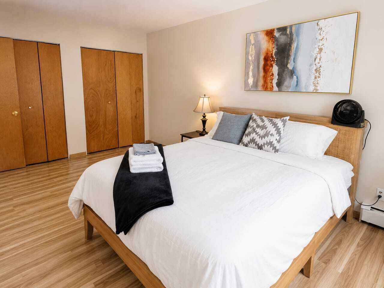 A hotel room with a bed, a nightstand, a lamp, a fan, and a painting on the wall.